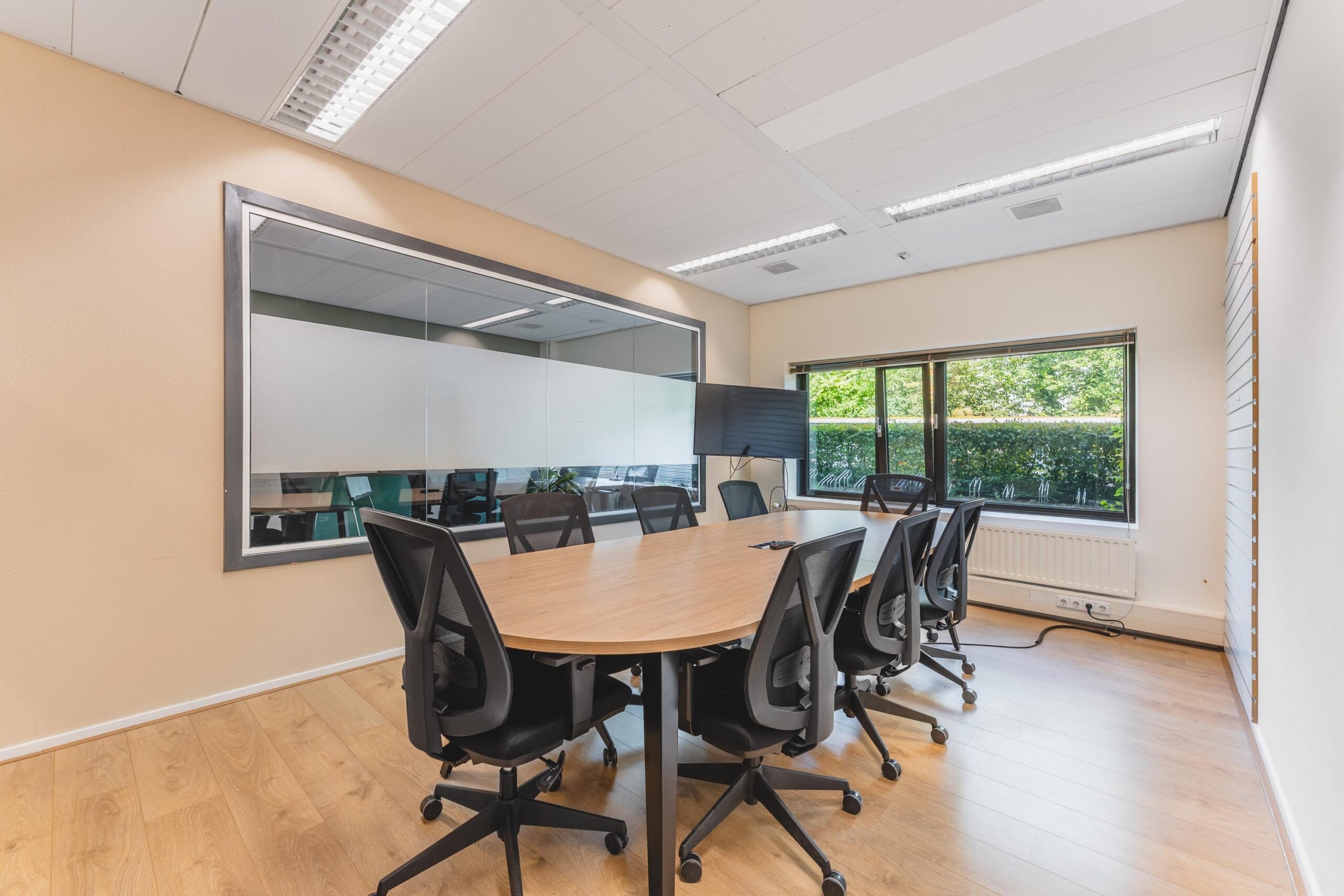 Vergaderruimte met ovale houten tafel, acht zwarte bureaustoelen, groot raam en tv-scherm aan de Prof. W.H. Keesomlaan.